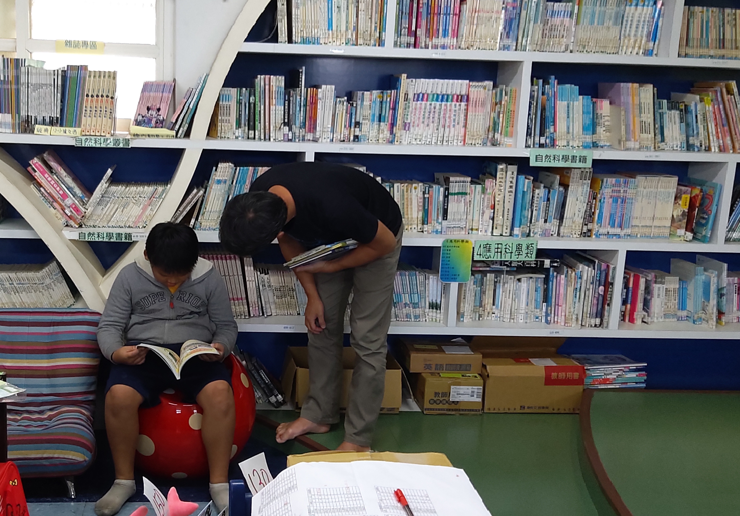 圖書館內，一名學生坐在紅色椅子上閱讀書籍，另一名學生站在旁邊查看書架。背景為分類整理的書籍，書架上標有科學和應用科學等標籤。地上放有筆記本和文具，呈現學習研究的氛圍。