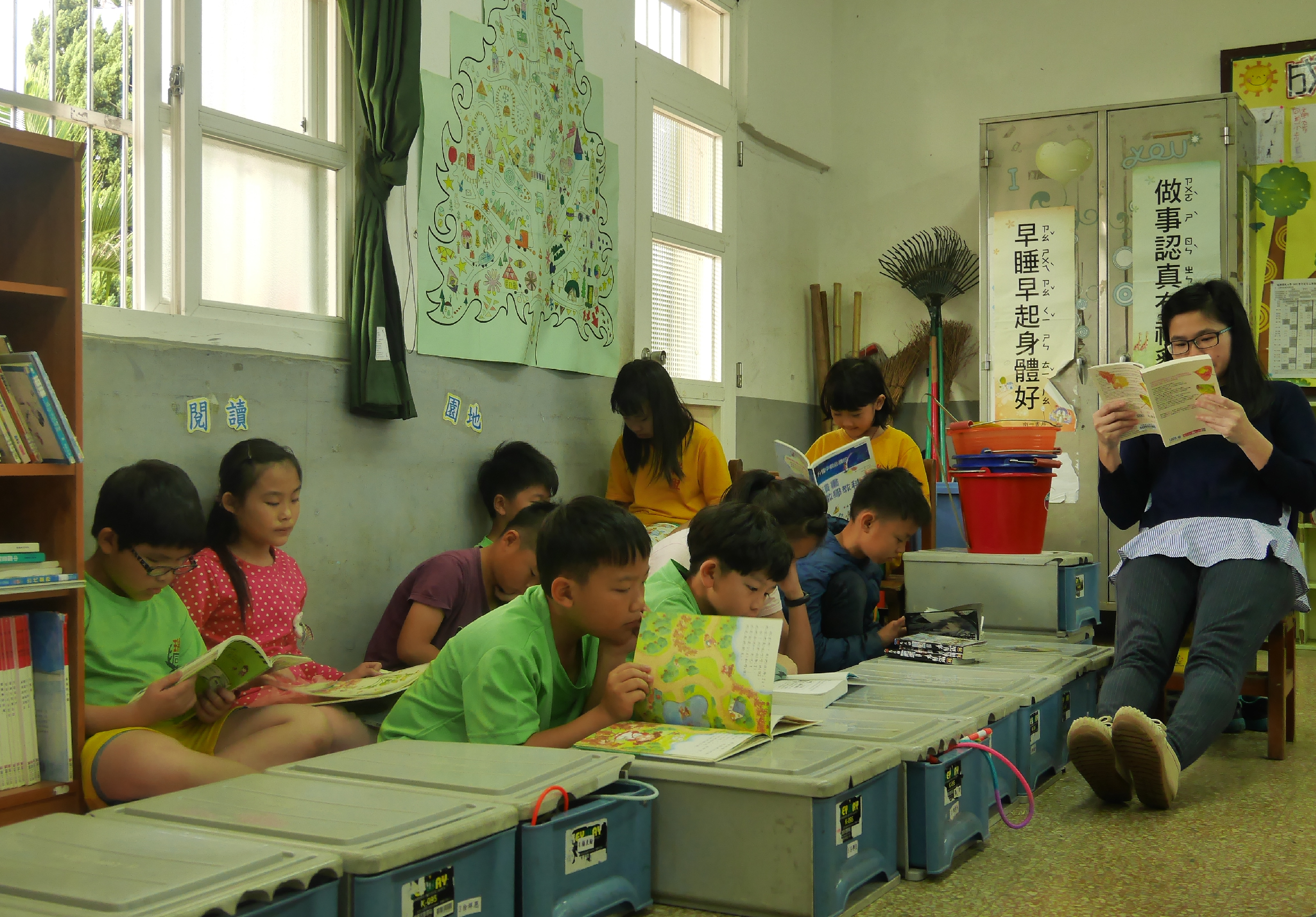 小學教室內，一位女性教師坐在地上向約十名學童朗讀繪本。孩子們圍坐在藍色儲物箱周圍認真聆聽。教室牆面為淡綠色，掛有中文標語和插畫。窗戶透入自然光，室內擺放書籍和教材。