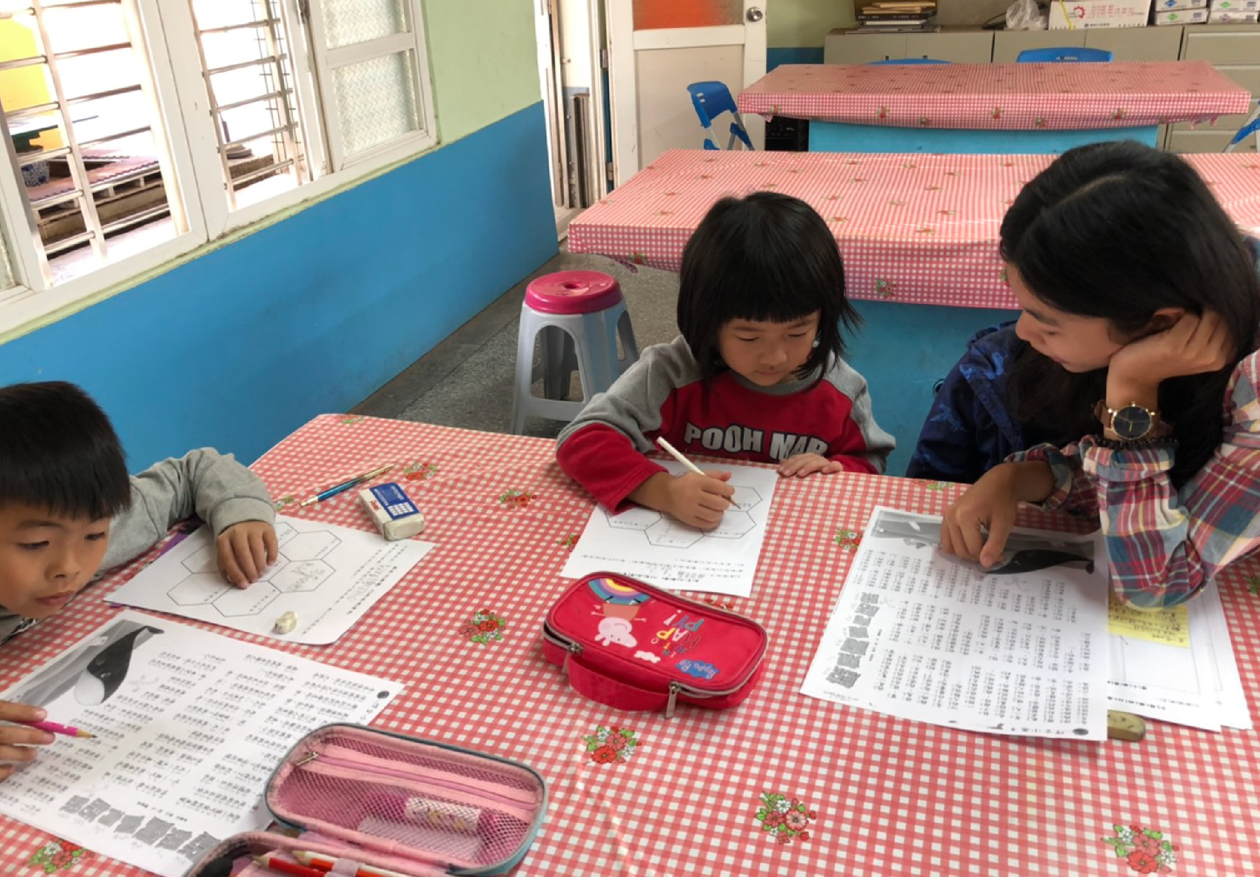 幼兒園教室內，四名年幼兒童圍坐在粉紅白格紋桌前，專注於功課練習。桌上擺放著練習卷、鉛筆盒和文具。教室牆面為藍白相間，背景可見粉紅色睡墊。一名女性教師在旁協助指導。