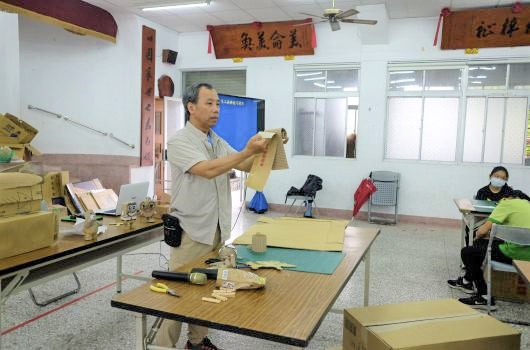 傳統手工藝教室中，男性講師在工作台前展示布料製作技巧，學生坐在一旁觀看。教室牆上掛著中文書法，擺放著各式手工藝材料和工具，呈現文化傳承的教學場景。