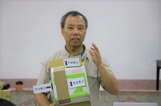 一位中年男性在室內進行產品展示演講，手持竹製建材樣本板，板上標示著不同材質和顏色的竹製品樣本。他穿著淺色上衣，右手做出説明手勢，背景為白色牆面，桌上放有黃色物品。