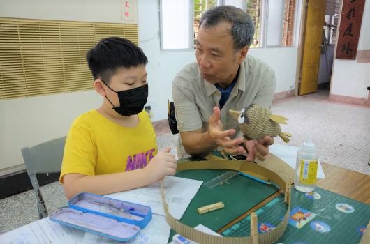 一名戴著黑色口罩、穿著黃色上衣的男孩坐在桌邊，與一位中年男性講師一起進行生物教學活動。講師正拿著一隻蜥蜴，向男孩展示和解說。桌上擺放著學習教材、工具箱和觀察記錄卡。室內環境為教室設置。