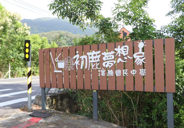 木製棕色招牌立於道路旁，上面寫著「初心夢想家」和「香島國民中學」，背景為青山綠樹，左側可見黃黑相間的安全警示柱和紅綠燈。
