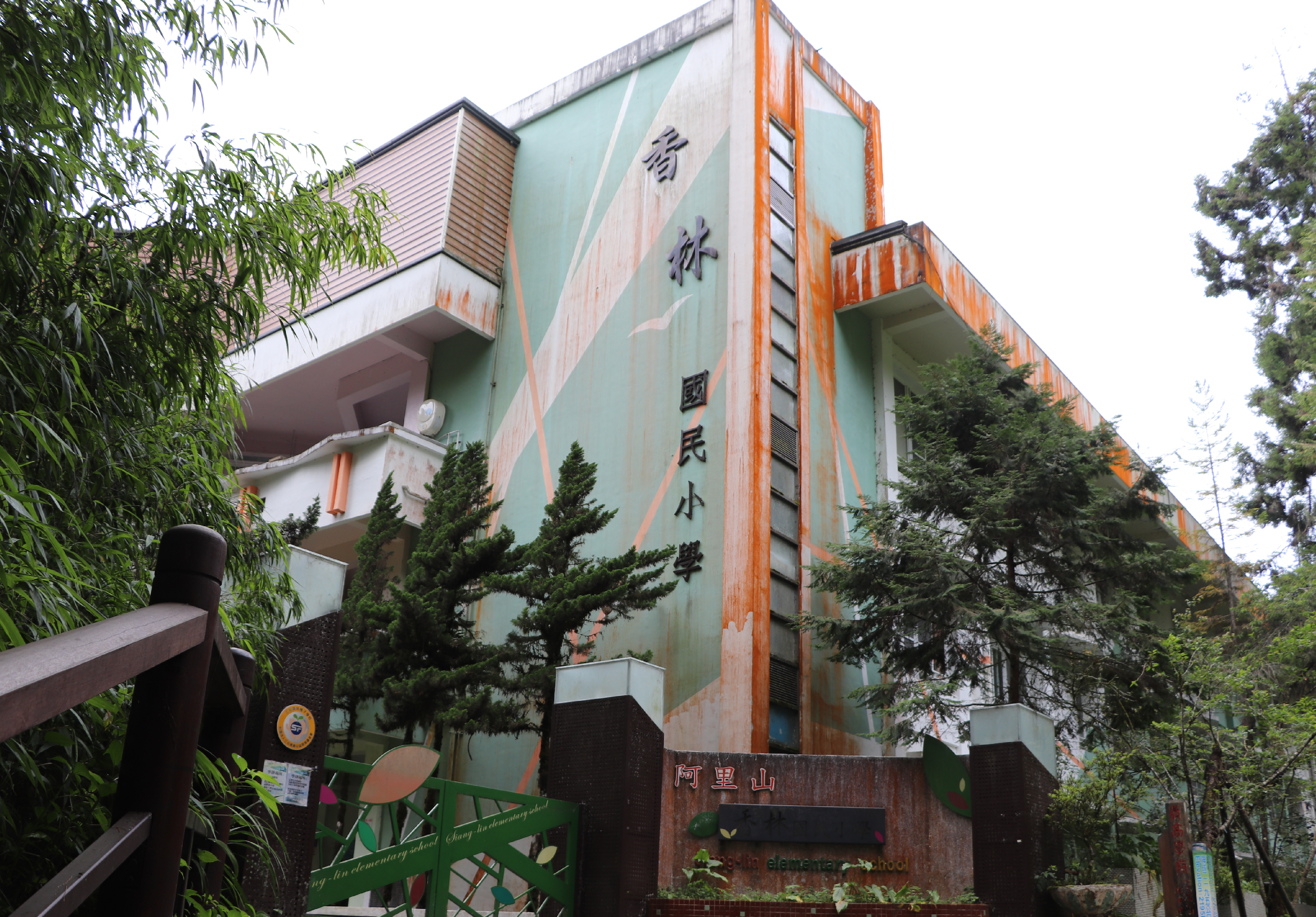 阿里山國民小學建築物外觀，淺綠色牆面配紅褐色鐵皮，牆上書寫中文校名。建築周圍種植綠樹和竹子，校門口立有棕色木製標誌牌。建物呈現歲月痕跡，具有山區校園特色。