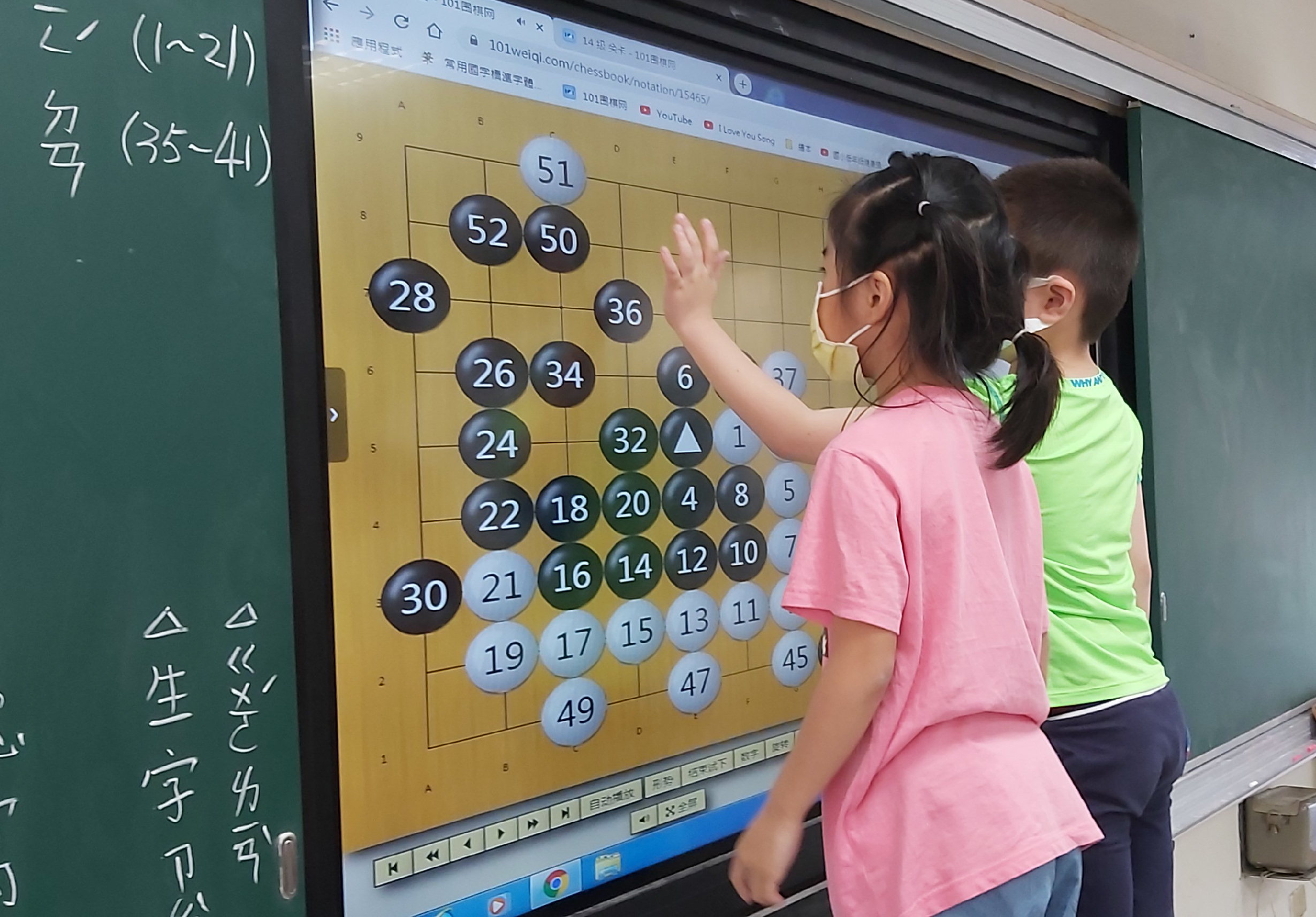 課堂上，兩名學生在互動式電子白板前學習圍棋。螢幕顯示棋盤及編號棋子，左側黑板寫有中文字。學生穿著粉紅色和綠色上衣，正指向棋盤進行互動學習。