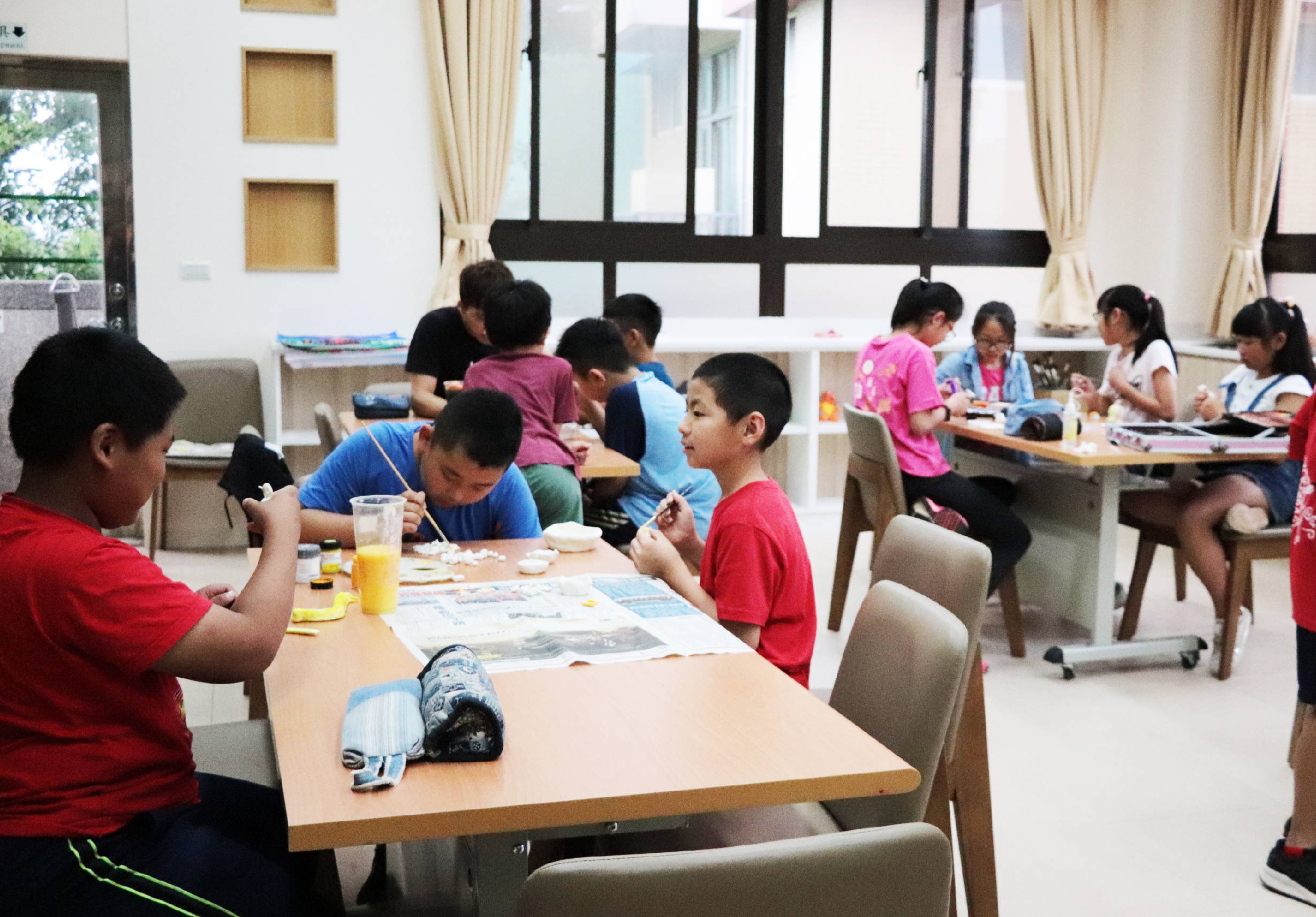 明亮的室內教室，多名兒童坐在數張木製餐桌前進餐和繪畫。前景兒童穿著紅色上衣正在用餐，中景兒童在繪畫創作，背景有成人監督。窗戶採光良好，牆上有米色窗簾和木製壁櫃。