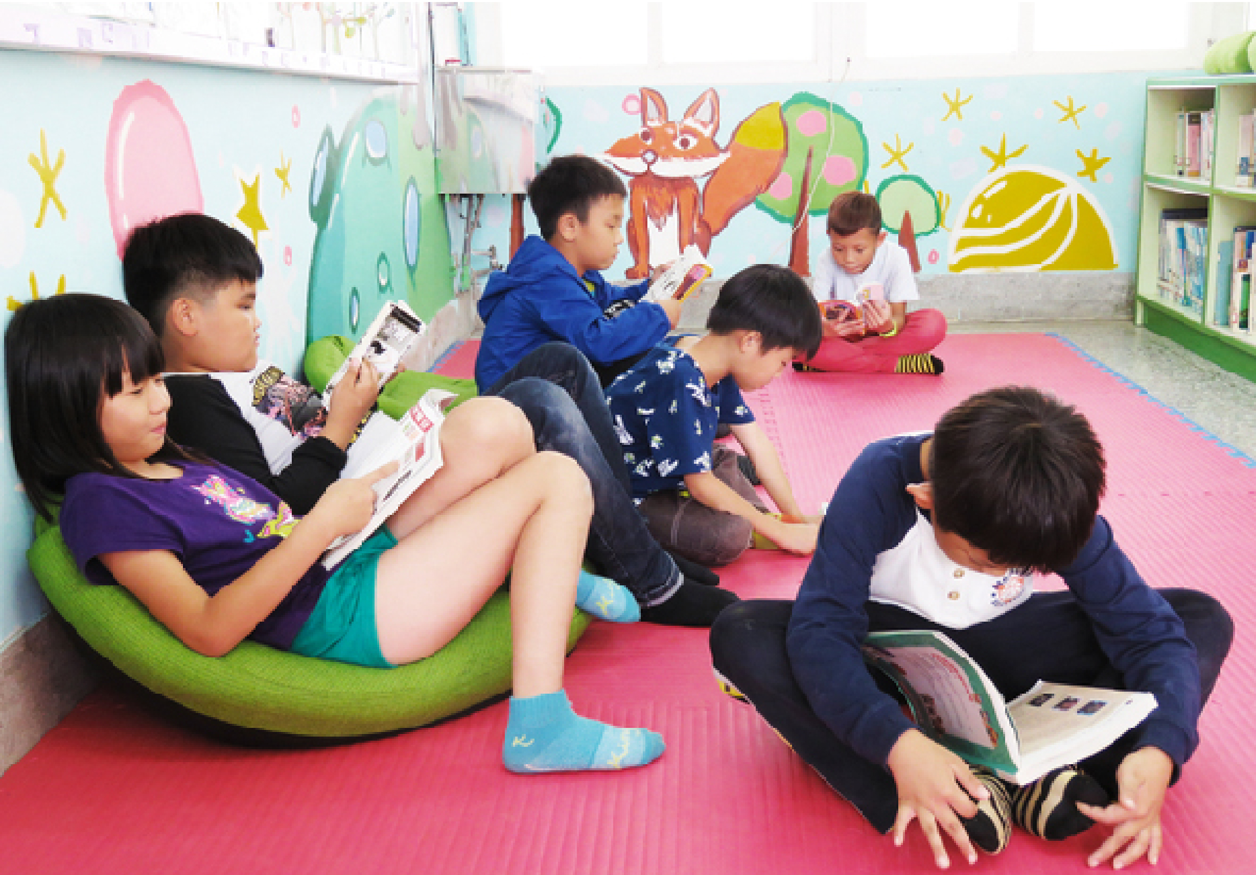 一間彩色兒童教室內，六名小學生坐在粉紅色地墊上，圍成一圈閱讀繪本。其中一名女童坐在綠色豆袋椅上，其他孩童分散坐在地上。牆面裝飾著可愛的狐狸、植物和星星圖案。右側有綠色書櫃。這是一場溫馨的集體閱讀活動。