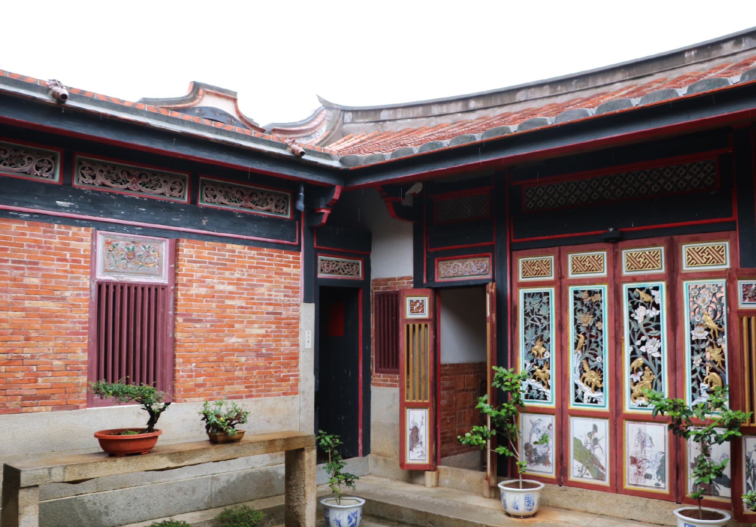 傳統中式庭院建築，紅磚牆面配黑色木質門框，翹角瓦頂。庭院內擺放陶土盆栽，右側牆面掛滿木刻裝飾板和古典山水畫，展現典雅的東方美學風格。