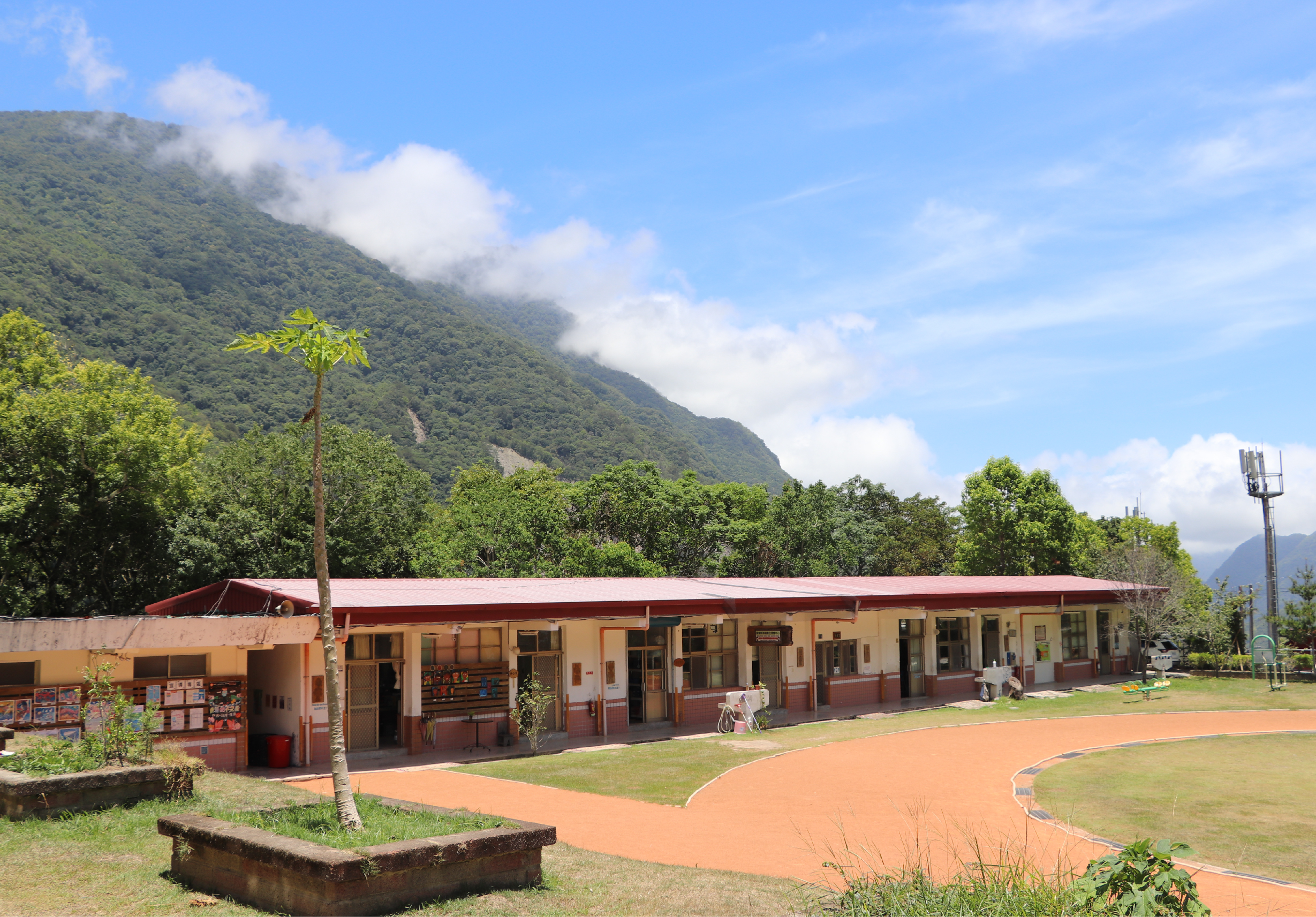 山腳下一所學校的校園景觀，建築物為紅白相間的平房設計，前方有橘紅色的跑道和綠色草坪。遠處青山綠林，白雲飄浮，右側立有通訊塔。