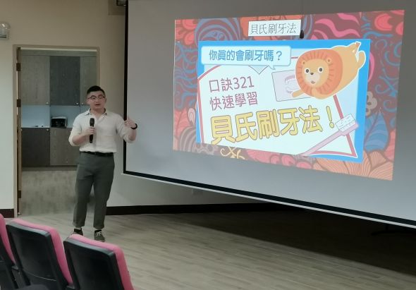 一位男性講者在教室進行牙齒刷牙法的教學演講。身後投影幕上展示中文標題「直式刷牙法」和「口訣321快速學習」，搭配卡通獅子角色圖案，介紹正確的刷牙技巧。