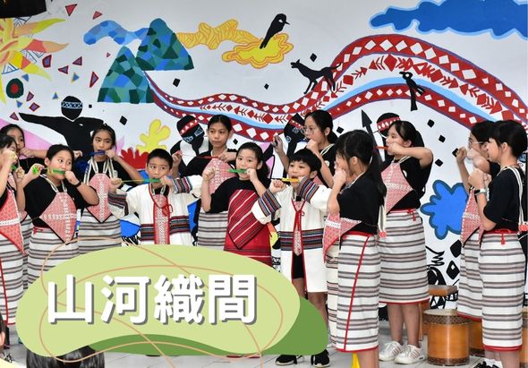 一群穿著紅白條紋服裝的兒童在舞台上表演，背景有彩色圖案和紅色波浪裝飾。舞台下方有綠色標牌寫著「山河織間」。孩子們擺著各種姿勢，展現活力和歡樂的表演氛圍。