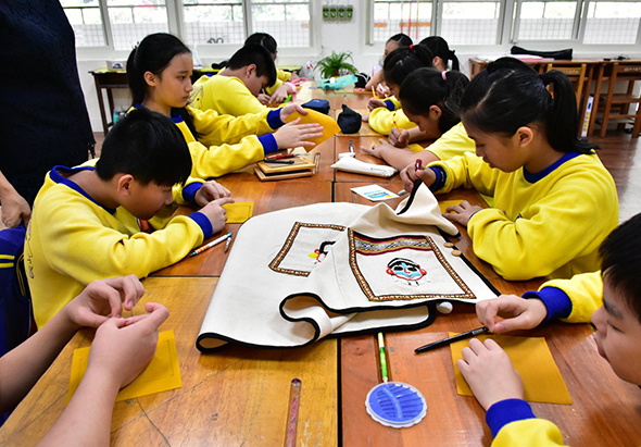 一群穿著黃色制服的學生坐在木製長桌邊，專注地進行手工藝活動。桌上擺放著白色紙板、彩色膠帶和製作材料。教室明亮寬敞，窗戶採光良好，學生們認真地製作手工藝品，呈現出充滿活力的課堂學習氛圍。