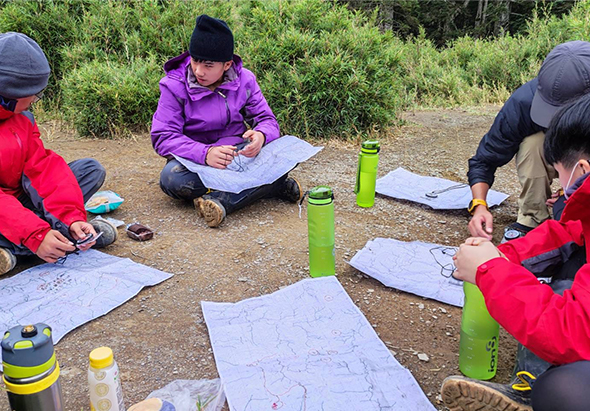 戶外地圖閱讀活動，四位穿著登山裝備的參與者圍坐在地上，各自查看地圖。地面放置綠色保溫瓶和指南針等工具，背景為灌木叢林，呈現登山或野外導航訓練的場景。