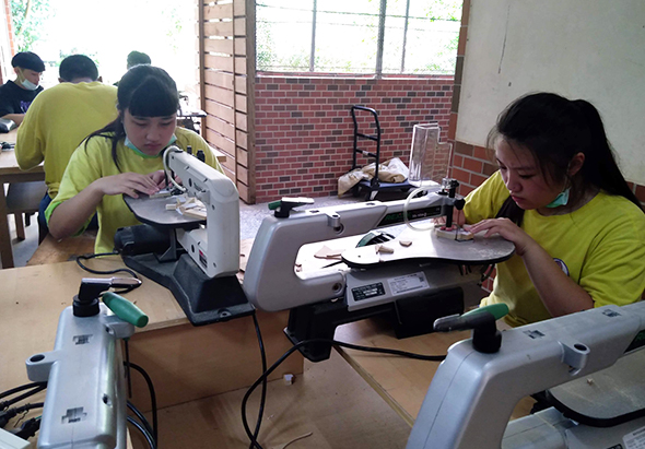 一間教室內，穿著黃綠色制服的學生坐在工作台前，操作電子設備進行組裝或檢測工作。桌上擺放著各式電子零件和工具，窗戶透入自然光，背景可見磚牆和其他學生在進行相似的實作活動。
