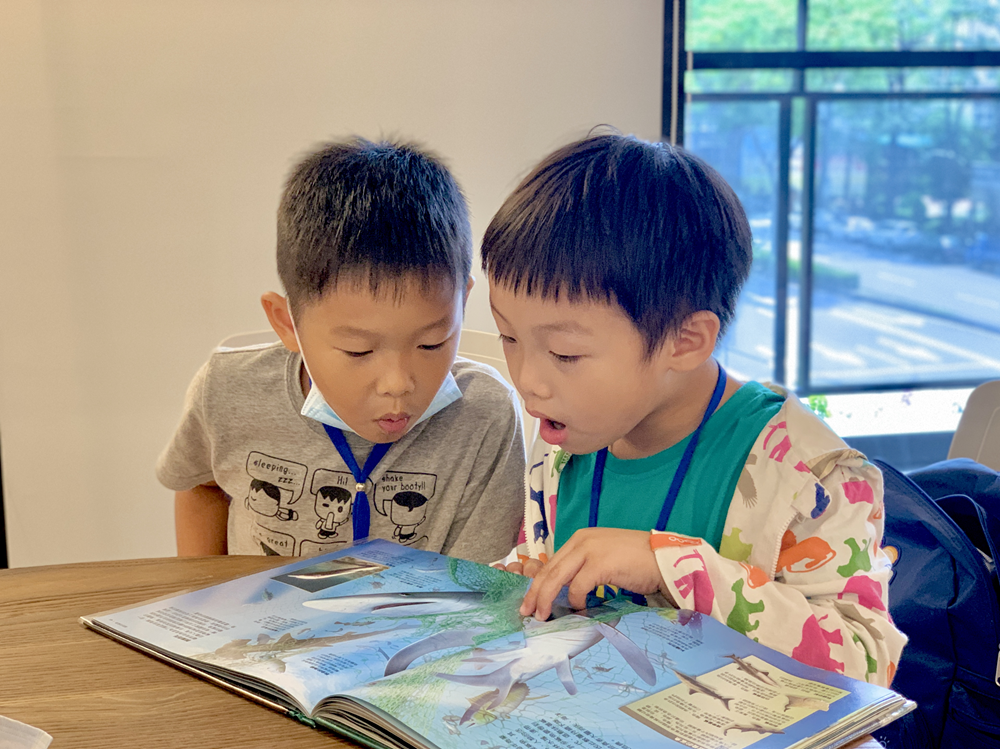 两名幼童在室内桌前一起阅读海洋生物主题的图书。左边穿灰色上衣的男孩，右边穿彩色衣服的男孩，两人都戴着识别证。明亮的落地窗在背景中，室外景色清晰可见。