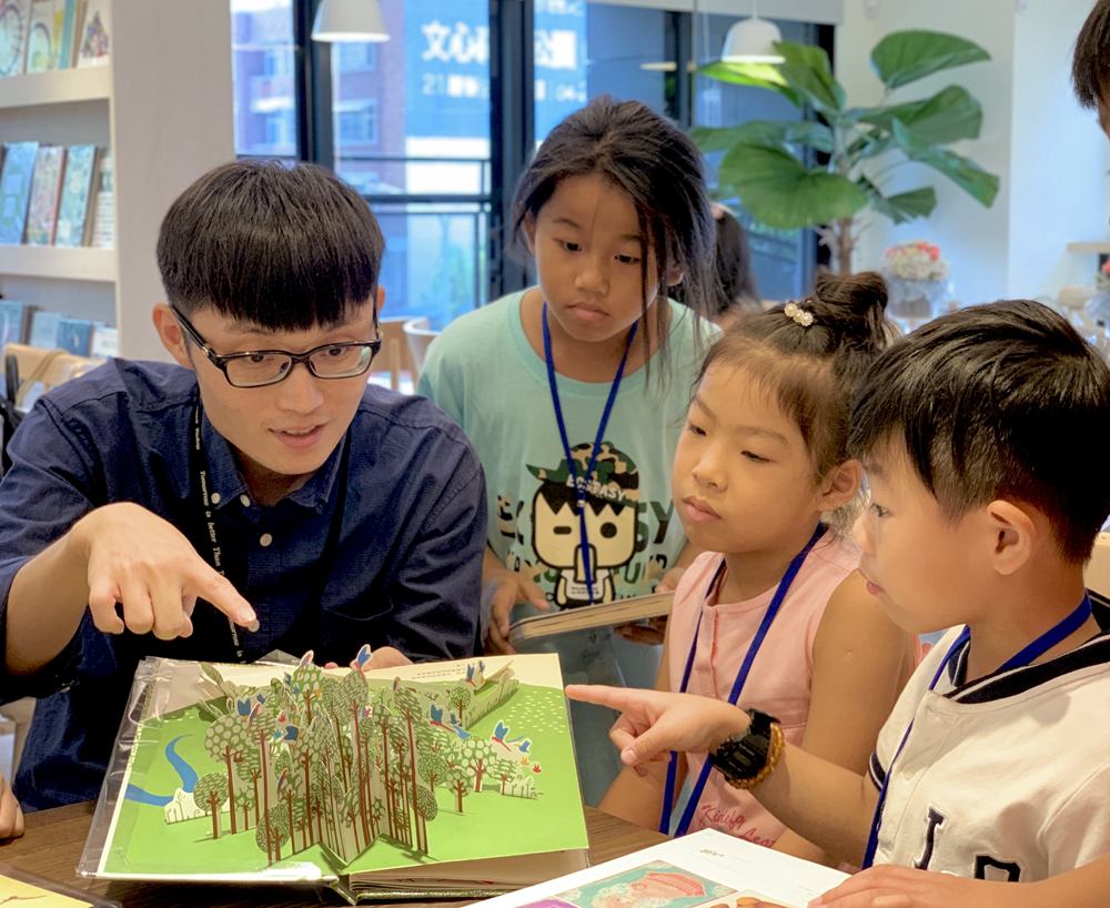 四名小學生在圖書館內圍繞一本打開的繪本，專注地觀看書中森林場景的立體插畫。左側男生穿著深藍色襯衫和眼鏡，其他學生穿著淺綠色和粉紅色衣服，都佩戴識別證。背景有書架、盆栽和落地窗。