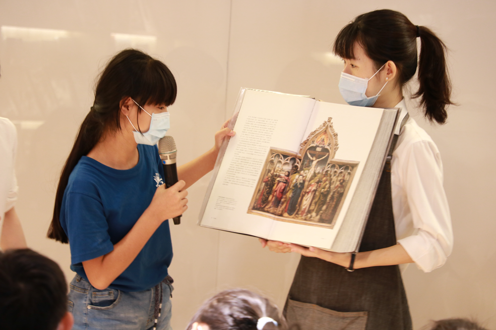 兩位戴口罩的女性在室內進行藝術展示講解，左方女性持麥克風，右方女性展示一本開啟的書籍，書頁上有宗教主題的古典繪畫作品。前方觀眾正聆聽講解。