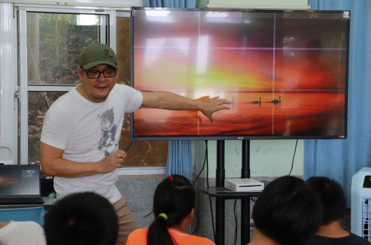 戴著綠色帽子和眼鏡的男性講師在教室內向學生展示大型液晶螢幕，螢幕上顯示紅色日落景象。數位學生坐在前排觀看演示，背景可見窗戶和藍色窗簾。