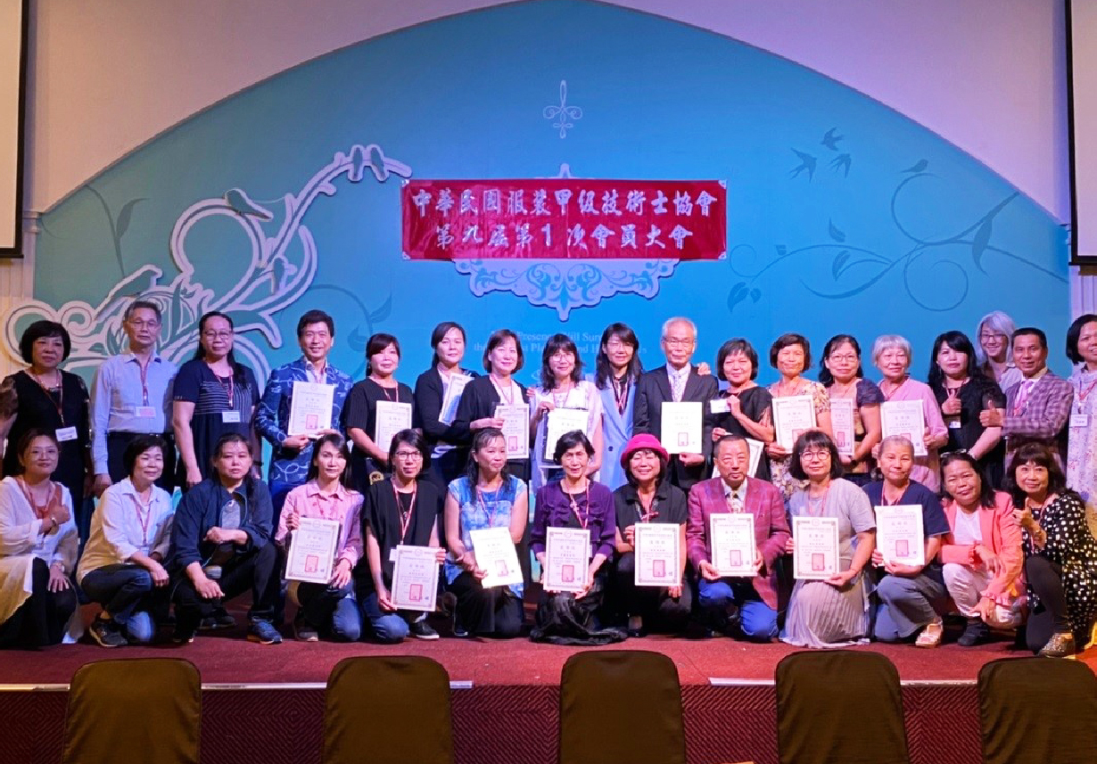 中華民國視覺藝術設計協會第九屆第一次會員大會舞台合影。兩排與會人士手持獲頒證書，背景為藍色裝飾幕牆及紅色會議標語。