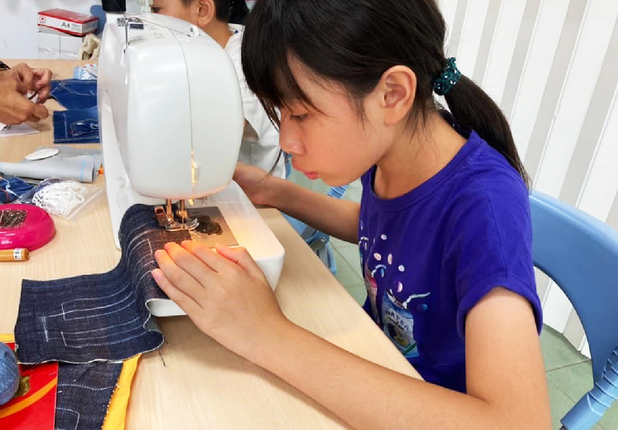 一位穿著紫色短袖上衣的女孩坐在木製工作台前，專注地使用白色縫紉機縫製牛仔布料。她的手指引導著深藍色牛仔布進入機器。工作台上散放著各色布料、剪刀和縫紉工具，背景可見其他學員在進行手工藝課程。