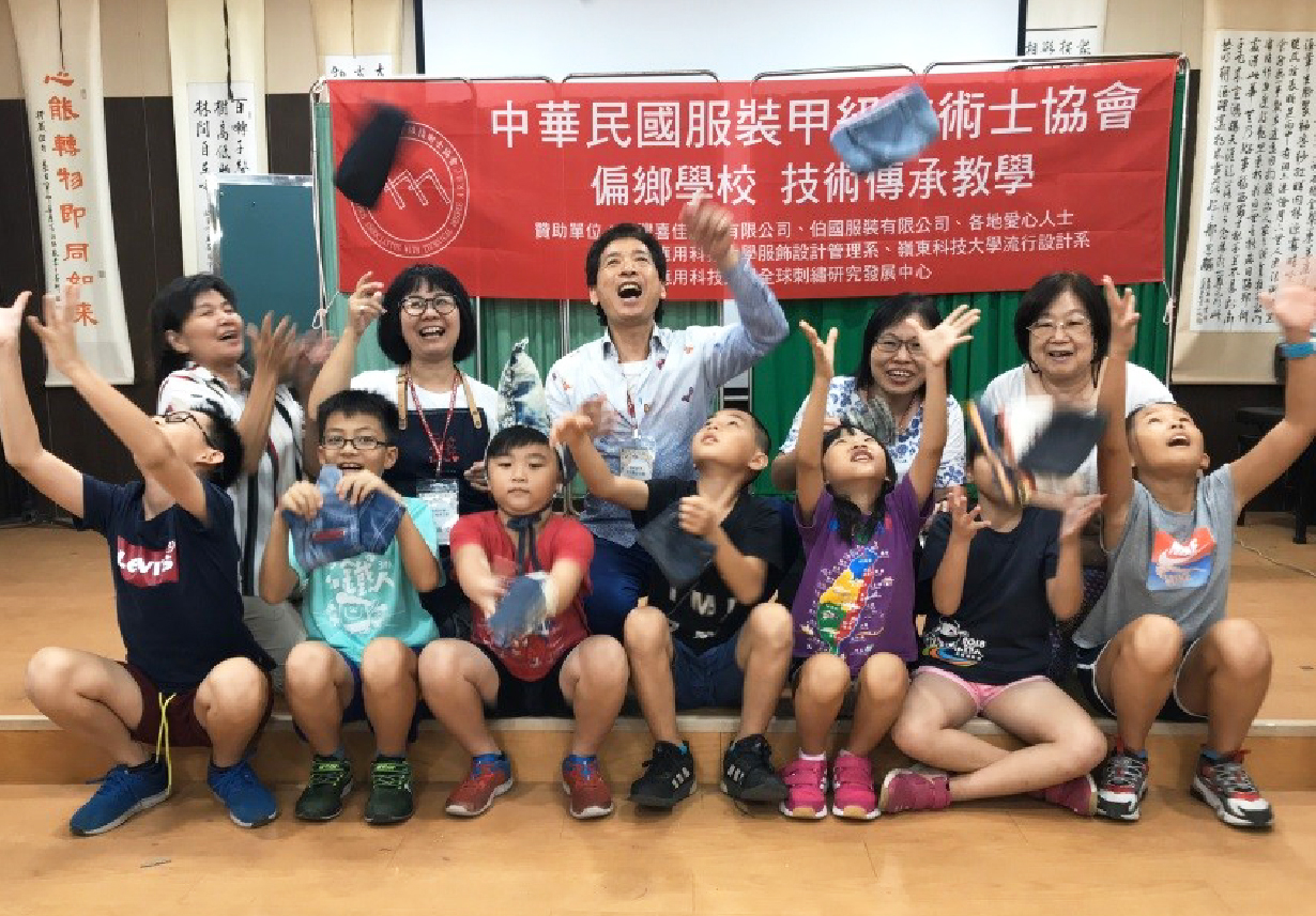 中華民國服裝甲級技士協會偏鄉學校技術博承教學活動現場，一群教師和學童在紅綠色布幕前歡樂地舉手慶祝，展現教學成果與互動的溫馨時刻。