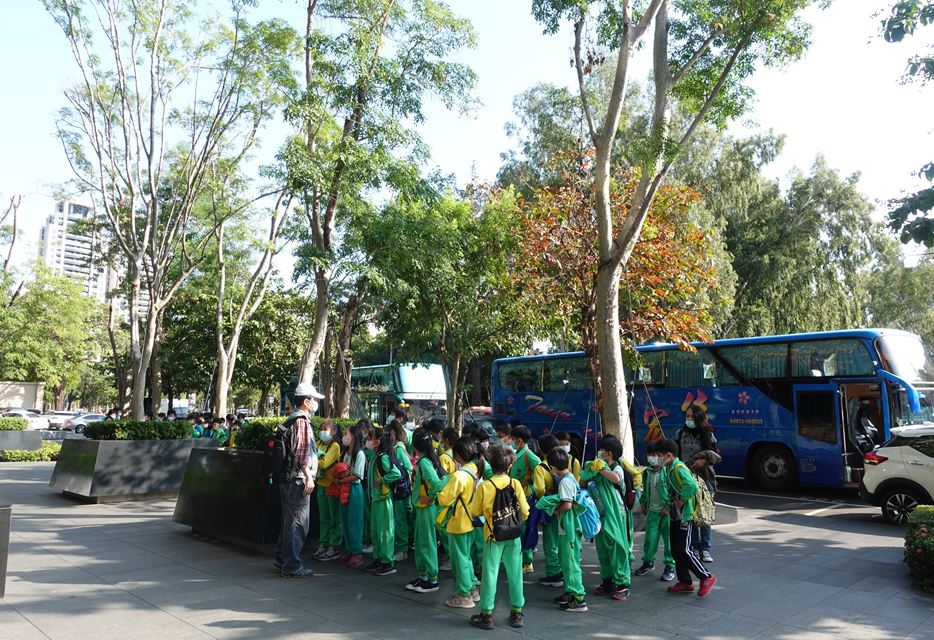 一群穿著綠色制服的學生在城市公園樹下集合，準備登上藍色大巴士。現場有多輛巴士停靠，高大的樹木提供遮蔭，背景可見高樓建築。這是一場學校校外教學或集體出遊活動。