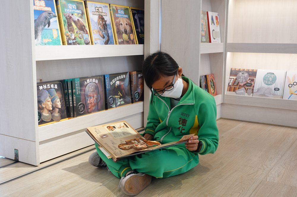 穿著綠色校服的小男孩坐在地上，戴著眼鏡和口罩，認真閱讀一本圖書。身後是白色書架，展示著多本中文書籍和教育類別的叢書。男孩專注於手中的書籍，呈現出專心閱讀的學習氛圍。