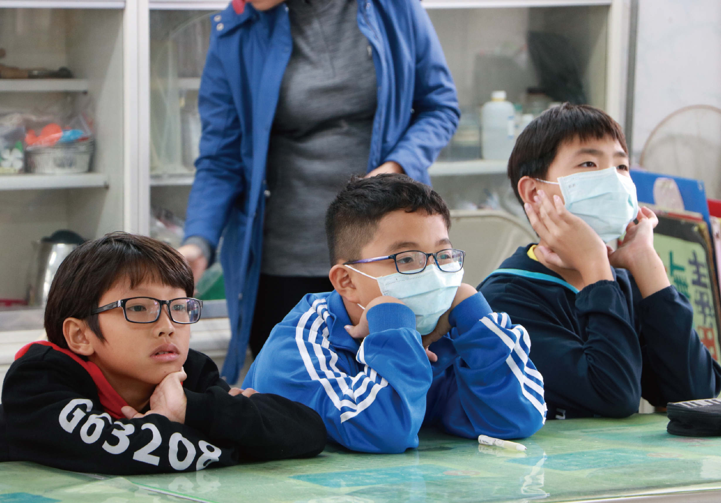 三名戴著口罩和眼鏡的學童躺在綠色地墊上，身穿藍色運動服。背景有一位成人穿著藍色外套站立，身後是白色儲物櫃，室內環境像是學校或教室。