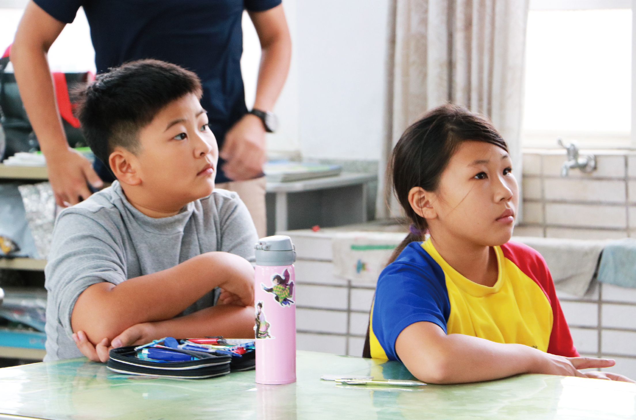 兩名小學生坐在教室課桌前專注地看向前方，左邊男童穿著灰色T恤拿著粉紅色水瓶，右邊女童穿著藍黃紅相間運動衣。桌上放有文具和書本，身後成人監督，背景為教室設施。