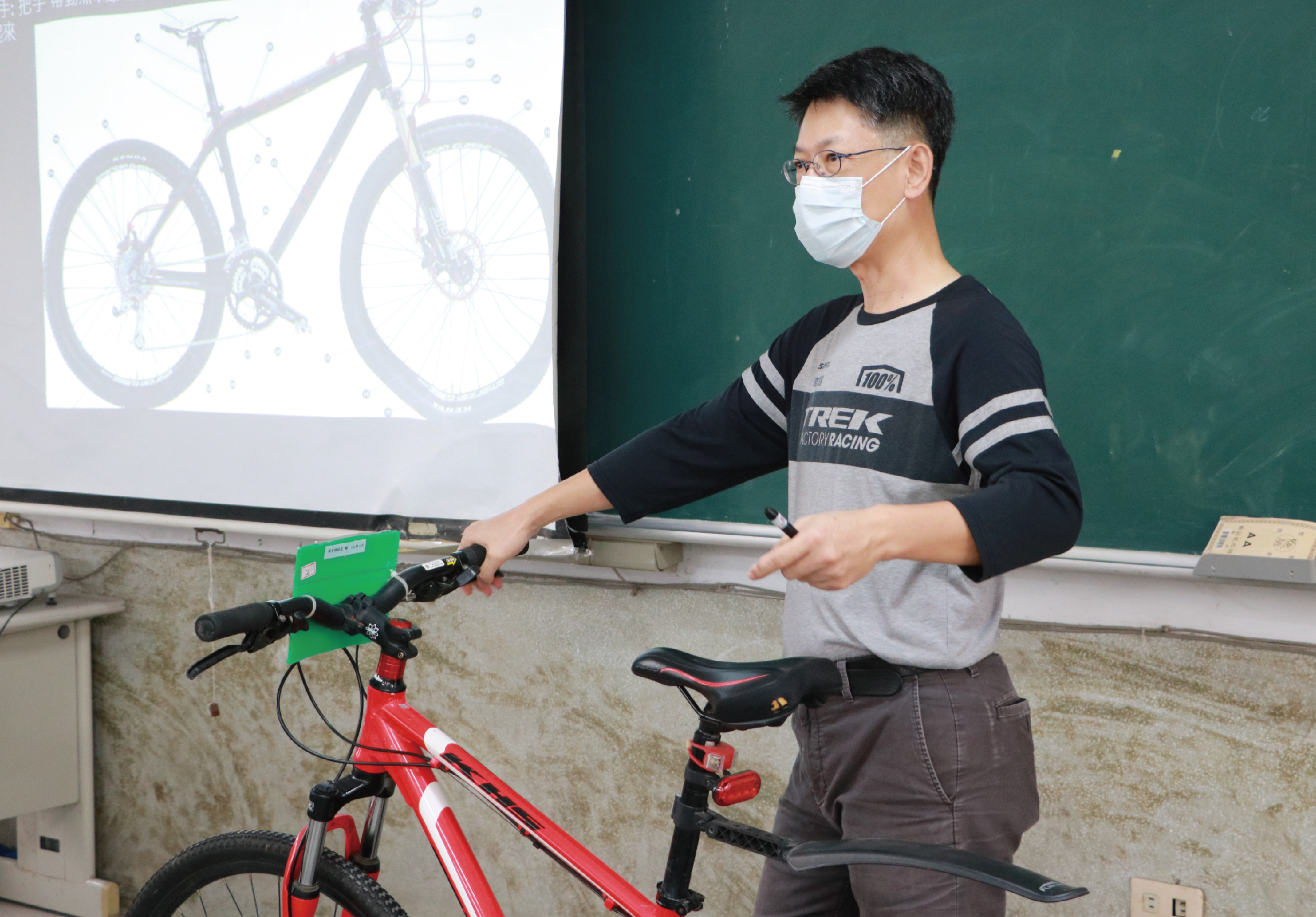 一位戴著口罩和眼鏡的男性講者在教室內進行自行車講座。他穿著Trek自行車隊服裝，正在展示一台紅色自行車。身後的螢幕投影著自行車的設計圖。綠色黑板和講台可見於背景。