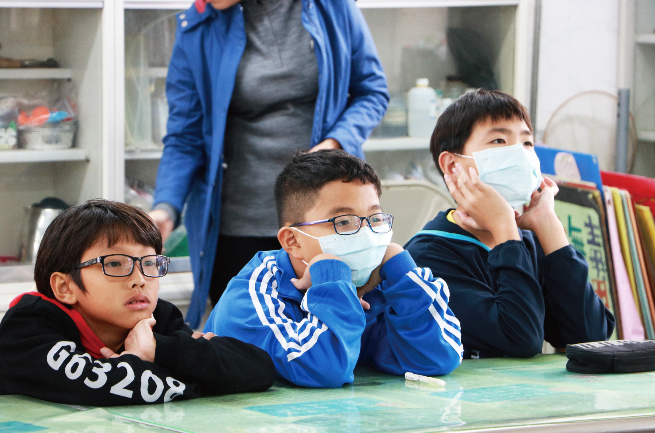 教室內，三名戴著口罩和眼鏡的小學男生坐在綠色地墊上看向前方，身後站著穿著藍色外套的女性監督者。白色櫃子上擺放著教室用品和彩色書籍，背景為室內教學環境。