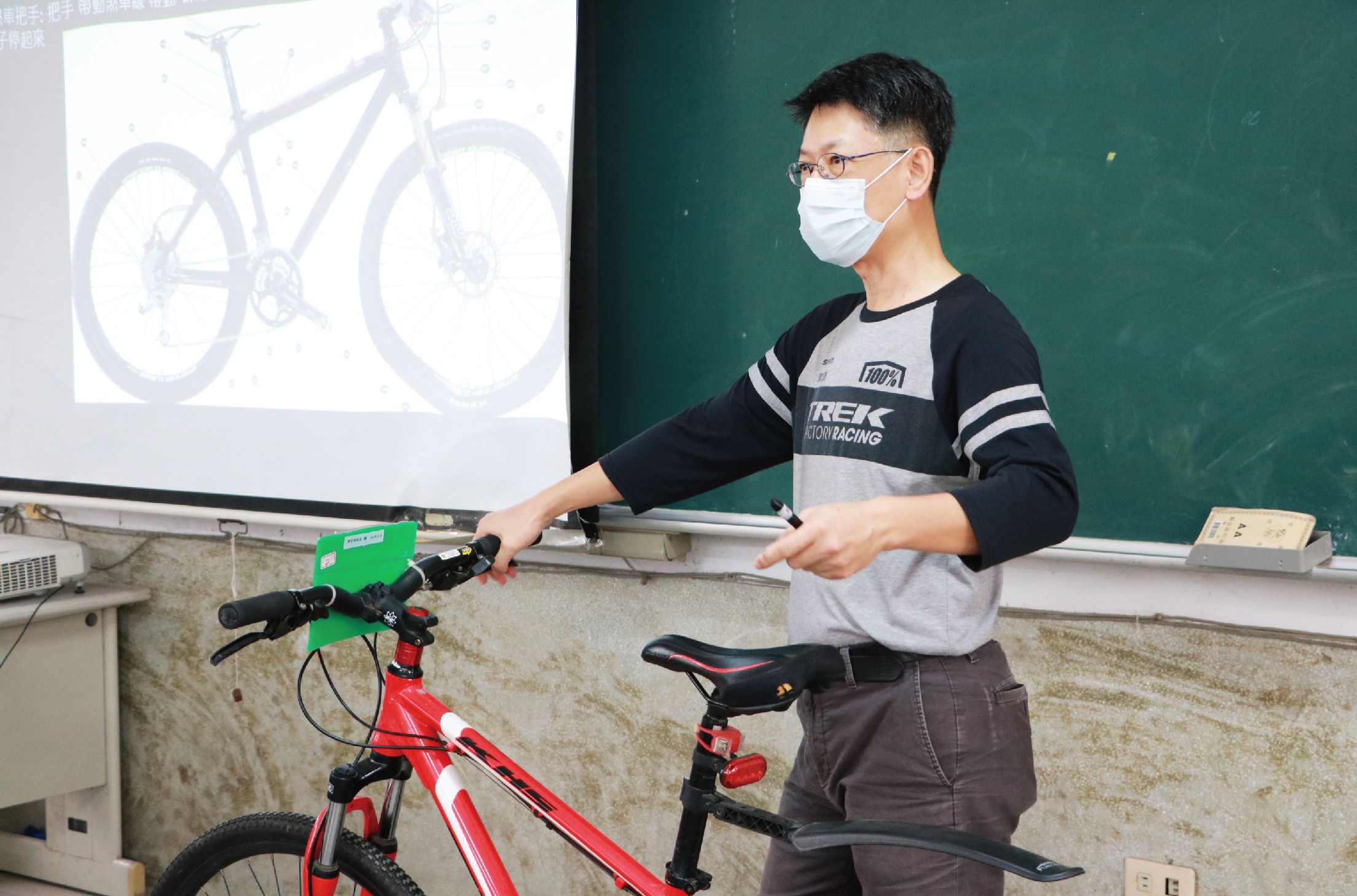 教室內，一位戴著口罩和眼鏡的男性講師正在展示一輛紅色自行車模型，身後投影螢幕顯示自行車的詳細圖片。講師穿著自行車相關品牌服飾，正向觀眾講解自行車的結構和功能。