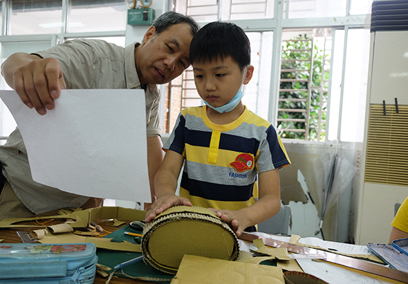一位成人和一名穿著黃黑條紋衣服的小男孩在室內工作坊進行手工藝活動。成人持著白色紙張指導，小男孩正在製作或裝飾一個黃色的圓形物體。桌上散放著各類製作工具和材料，背景可見窗戶和教室環境。