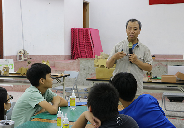 一名男性講師在教室前方拿著麥克風進行演講，三位學生坐在前排聆聽，桌上放著水瓶。教室白牆上掛著紅色布料，後方有粉紅色折疊椅。