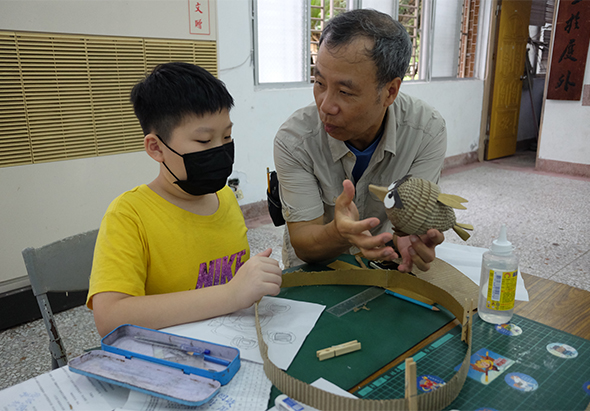 成人指導員與穿著黃色衣服、戴口罩的小男孩坐在教室桌邊，共同觀察和學習一條綠色的魚類標本。桌上放有學習工具、筆記本和水瓶，展示戶外教育或生物課程的互動學習場景。