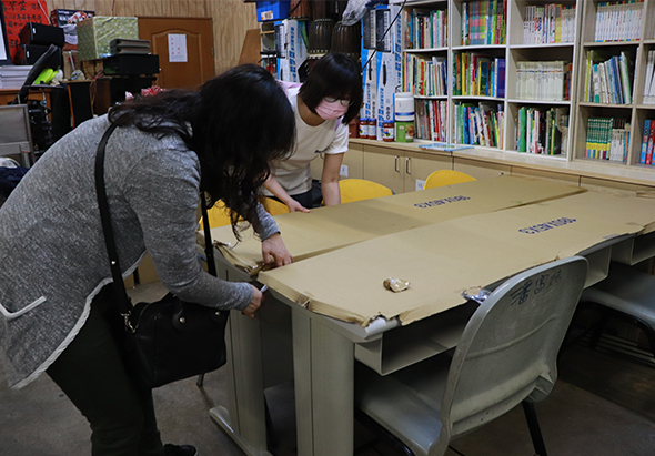 在學校圖書館內，兩位女性在米色桌子上進行活動。左側女性穿著灰色運動外套，右側女性穿著米色上衣，兩人正認真地在桌面上操作或組裝物品。背景可見書架和黃色椅子。
