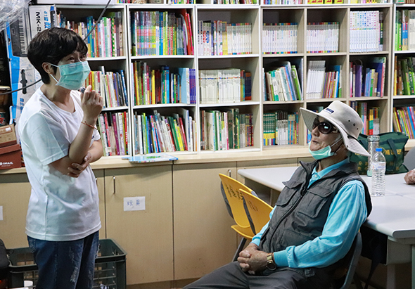 圖書館內，一位戴口罩的工作人員站立，與坐在黃色椅子上戴著帽子和墨鏡的訪客交談。背景是整面的書架，擺放著各類書籍。場景呈現友善的互動氛圍。