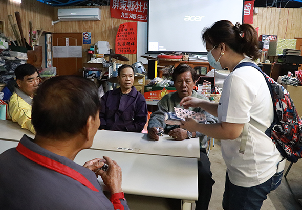 社區活動室內，一位年長婦女正向坐著的民眾展示手工藝品。室內木質裝潢，牆上貼有紅色標語和海報。約六位民眾聚集觀看，其中一人戴著口罩。背景可見投影幕、冷氣機和雜物，呈現溫馨的社區聚集場景。