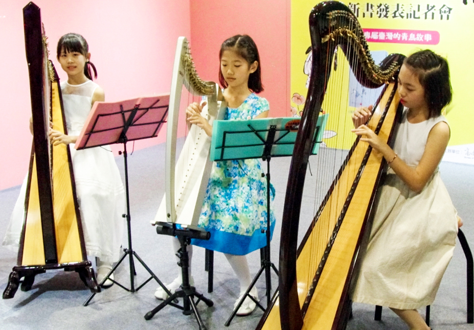 三位年輕女孩在室內演奏豎琴，每人持一把豎琴站在樂譜架前。背景為粉紅色和黃色牆面，展示中文文字和海報。這是一場音樂表演或教學示範活動。