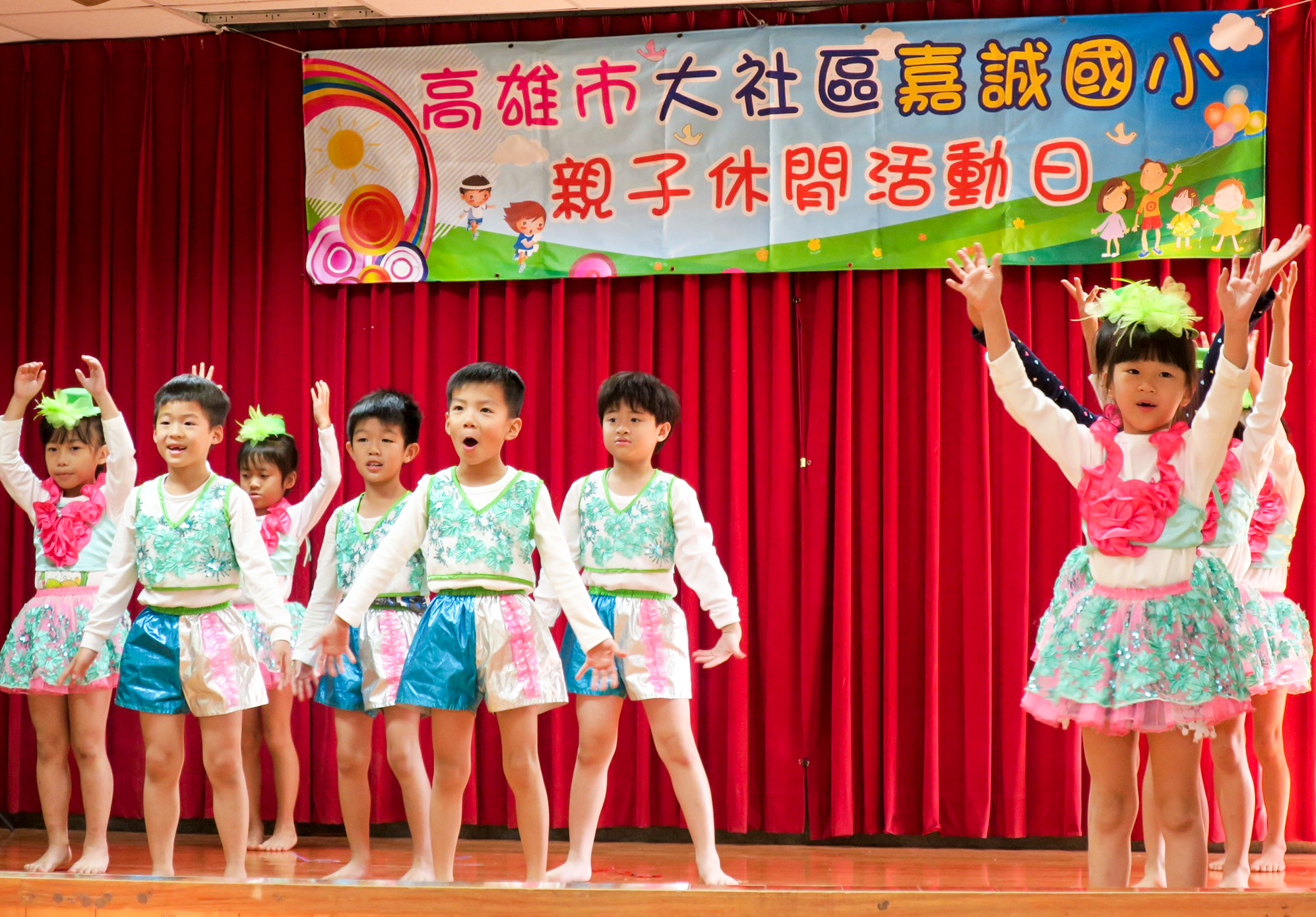 高雄市大社區嘉誼國小親子休閒活動日舞蹈表演，約十名穿著綠白粉紅花卉服裝的小學生在紅色舞台幕布前，做出歡樂舞蹈動作，舞台上方為彩色活動海報。