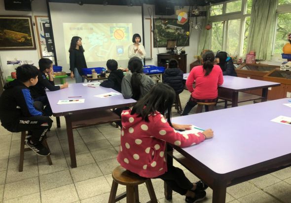 教室內學生坐在多張紫色和白色桌子前，面向講台。兩位女性教師站在投影幕前進行教學，螢幕上展示行星系統圖。教室窗戶透入自然光，牆上掛有教育海報。學生專注聽講，呈現互動式學習場景。