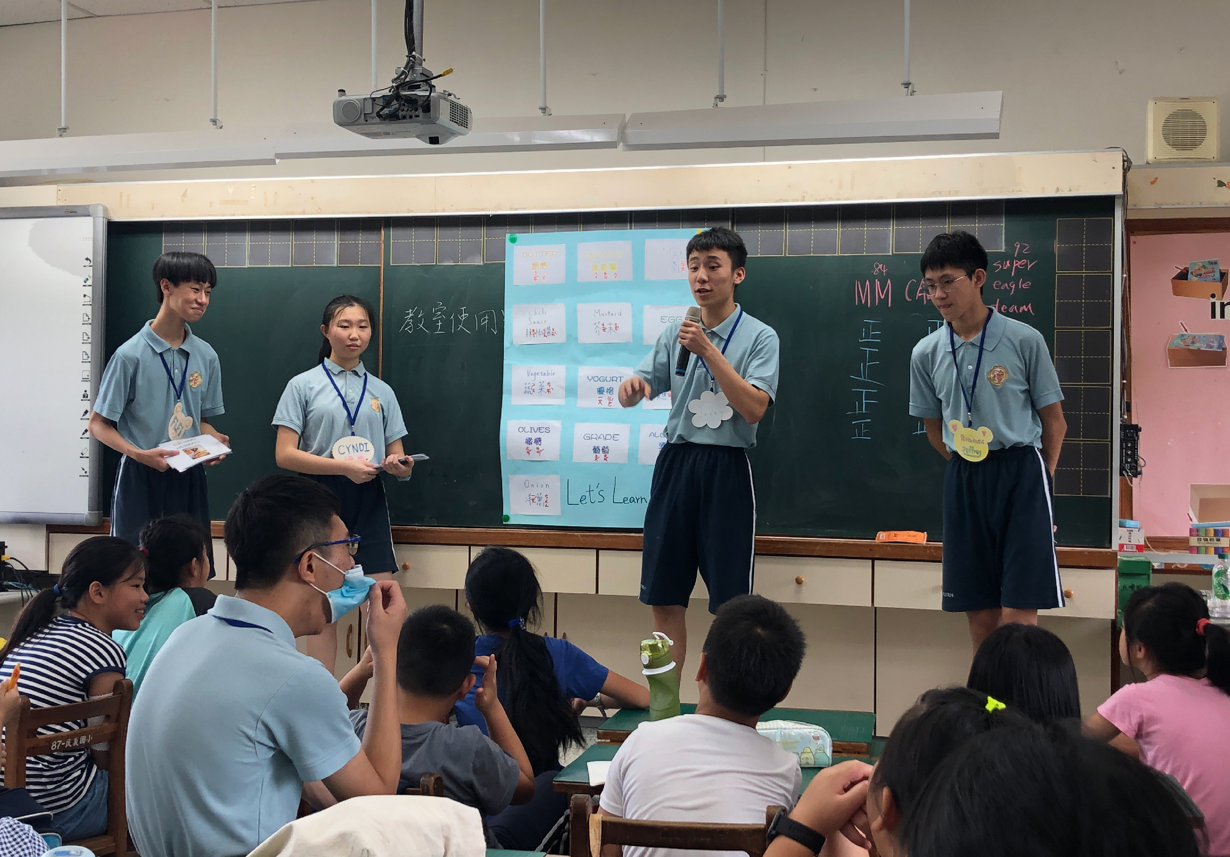 教室內，五位穿著制服的學生站在黑板前進行英語教學展示。黑板上貼有彩色便籤紙展示英文單字和短語。台下坐著約十位學生認真聆聽。現場使用投影機輔助教學，呈現互動式的英語學習活動。