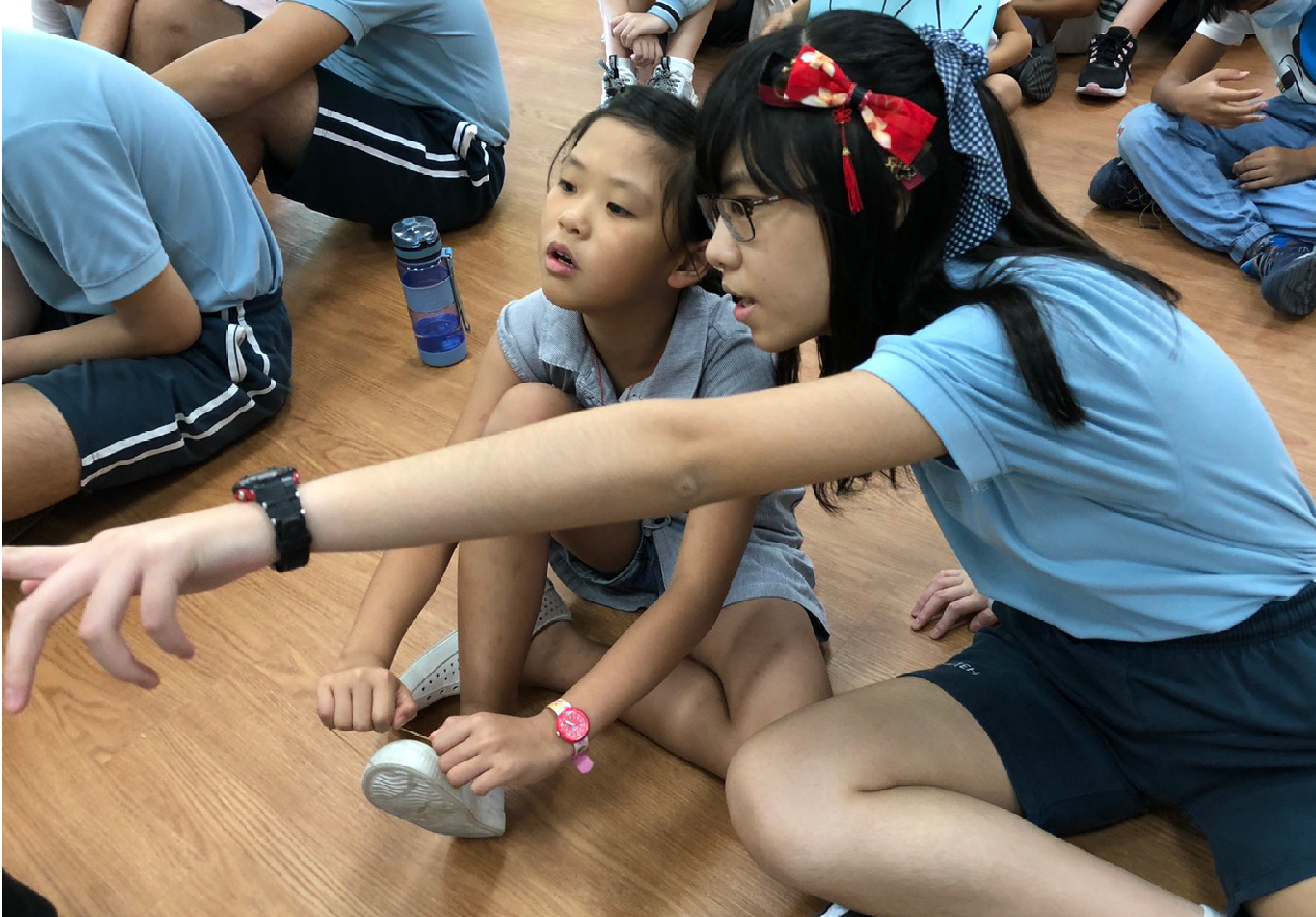 一群小學生圍坐在木質地板上，一位穿著藍色制服的女老師正向學生們演示或講解某樣物品。學生們認真聆聽，其中一名學生戴著紅色蝴蝶結髮飾。現場還有水瓶和其他學用品散置地上，呈現教室內的教學互動場景。