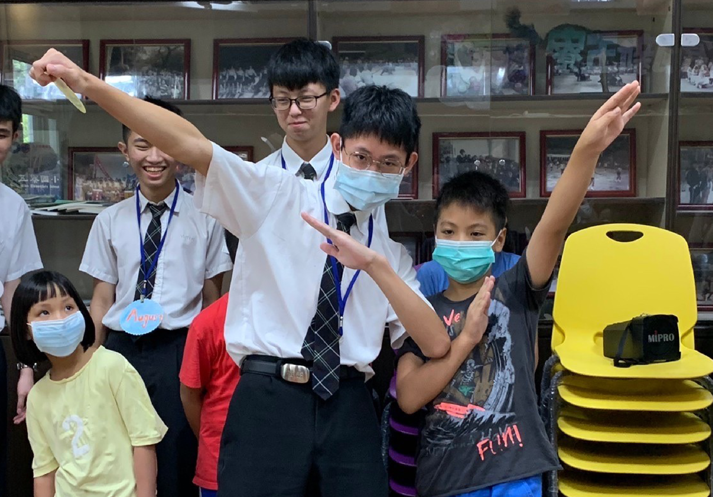 一群學生在室內舉辦活動，穿著校服的青少年和一名穿白衣的引導者展開雙臂做出歡樂的姿勢，多人戴著口罩，身旁有黃色的MIPRO音響設備。背景為展示歷史照片的玻璃櫃。