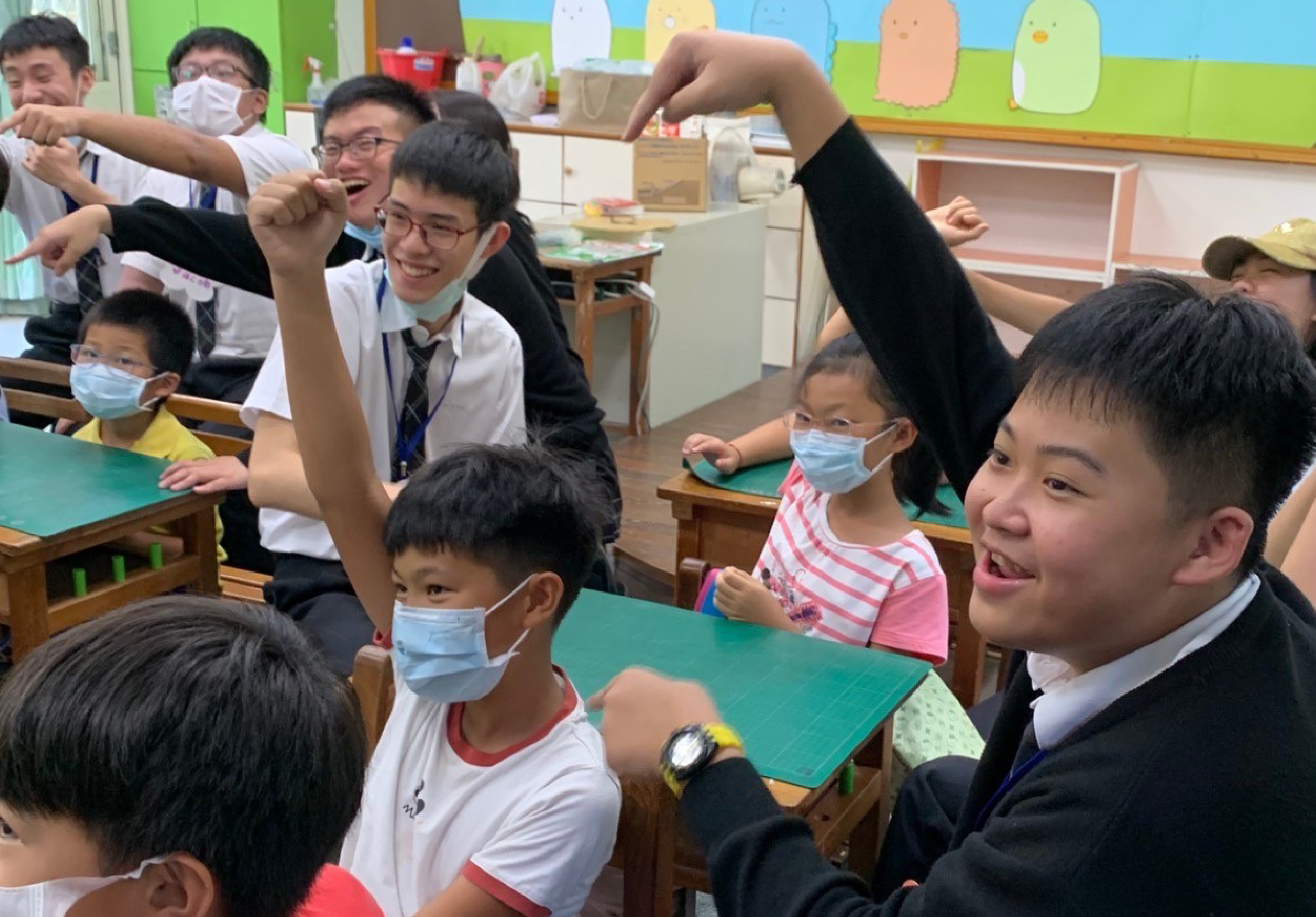 教室內，年輕教師與小學生們圍坐在綠色課桌旁，進行互動教學活動。學生們戴著口罩，舉起雙手開心地參與課堂，展現活潑的學習氛圍。背景可見彩色壁畫和教室布置。