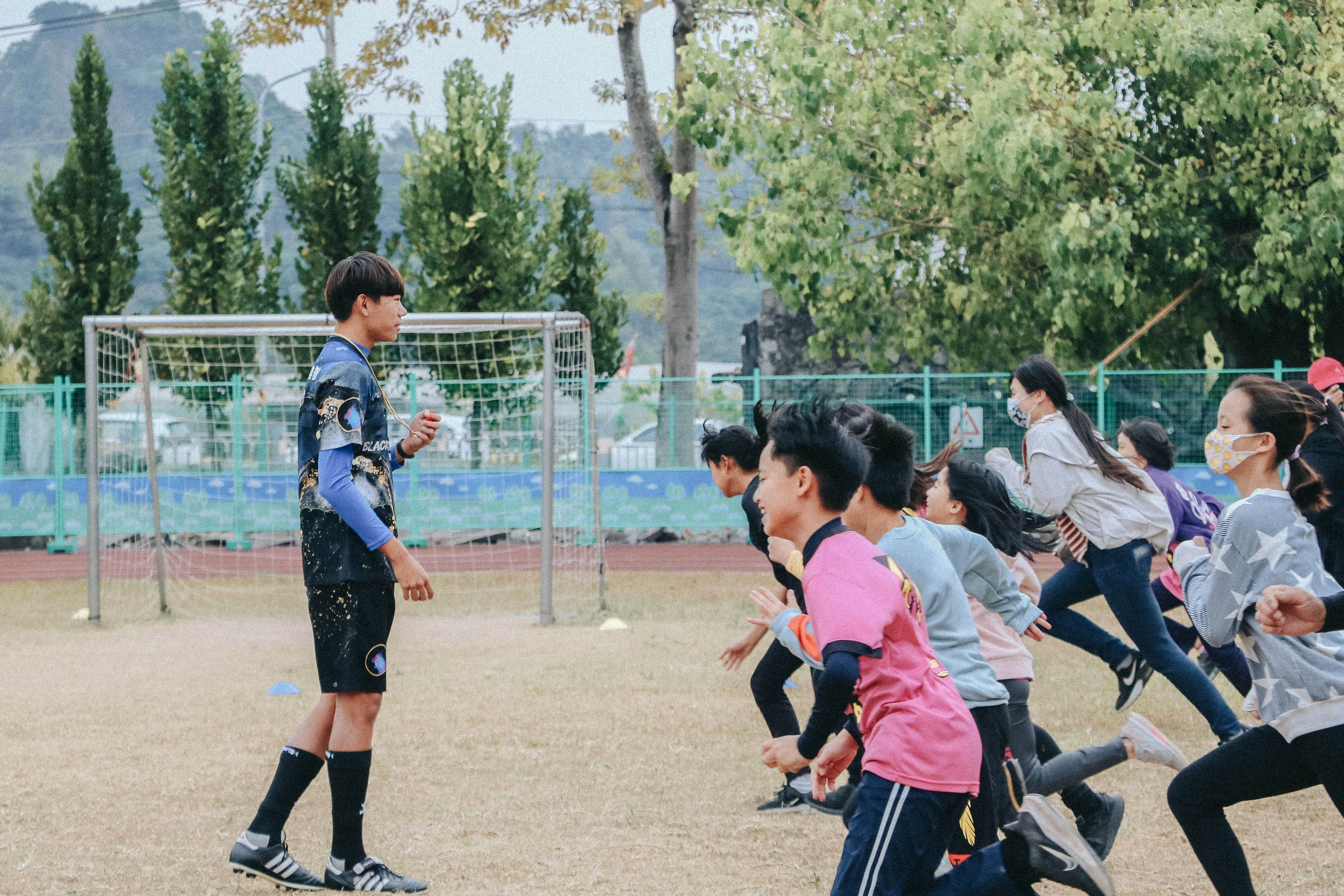 學校運動場上，一名穿著藍色運動服的教練或運動員正面向一群穿著各色運動服的學生講話指導。背景是足球門、綠色圍欄和翠綠的樹木，遠處可見山景。場景呈現體育課或訓練活動的進行。