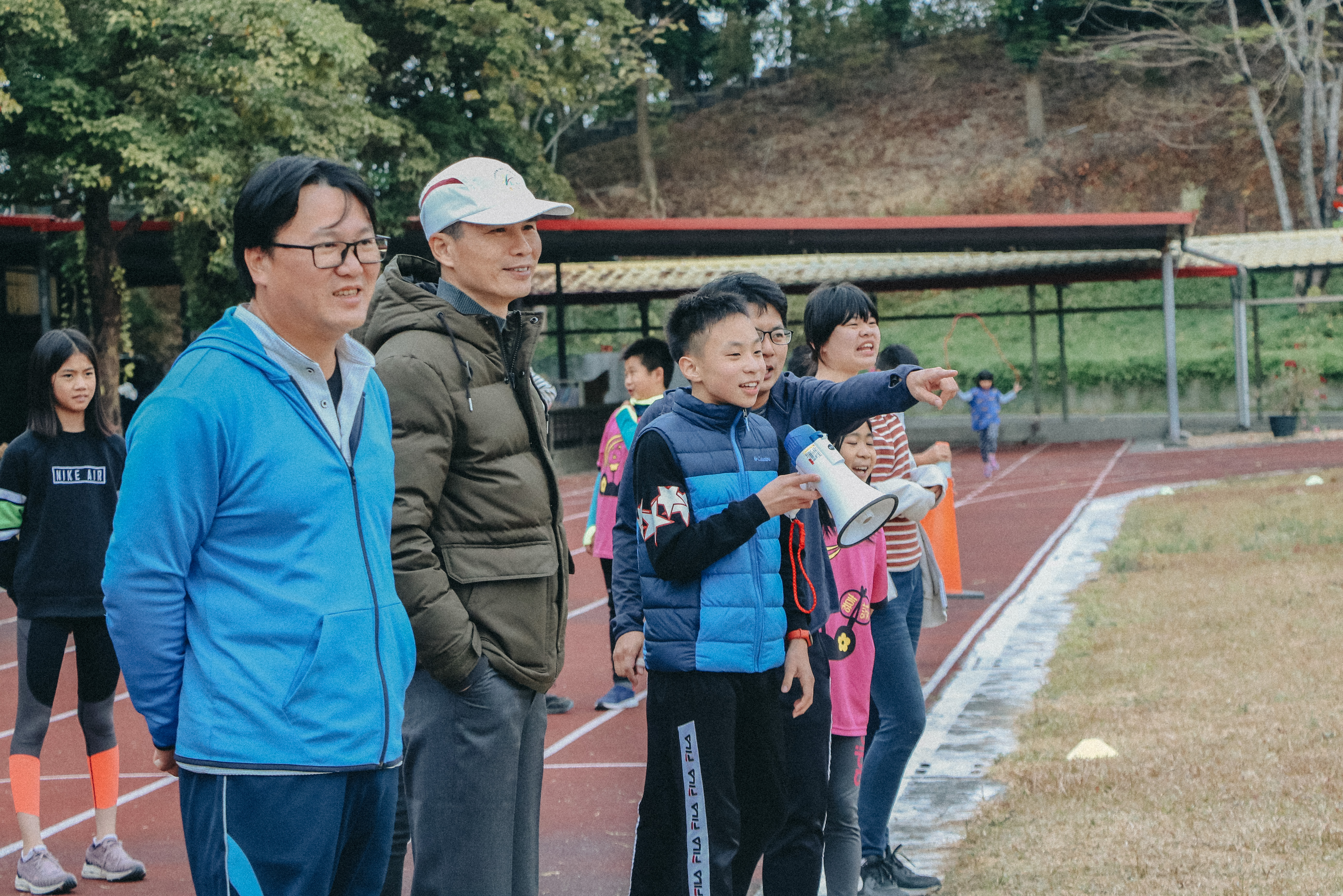 一群小學生在紅色跑道上排隊集合，一位穿著藍色夾克的男性成人和穿著棕色外套、白色帽子的成人站在前方帶隊。一名小男孩手持擴音器，其他孩子穿著各色運動服裝。背景為校園設施和樹木。