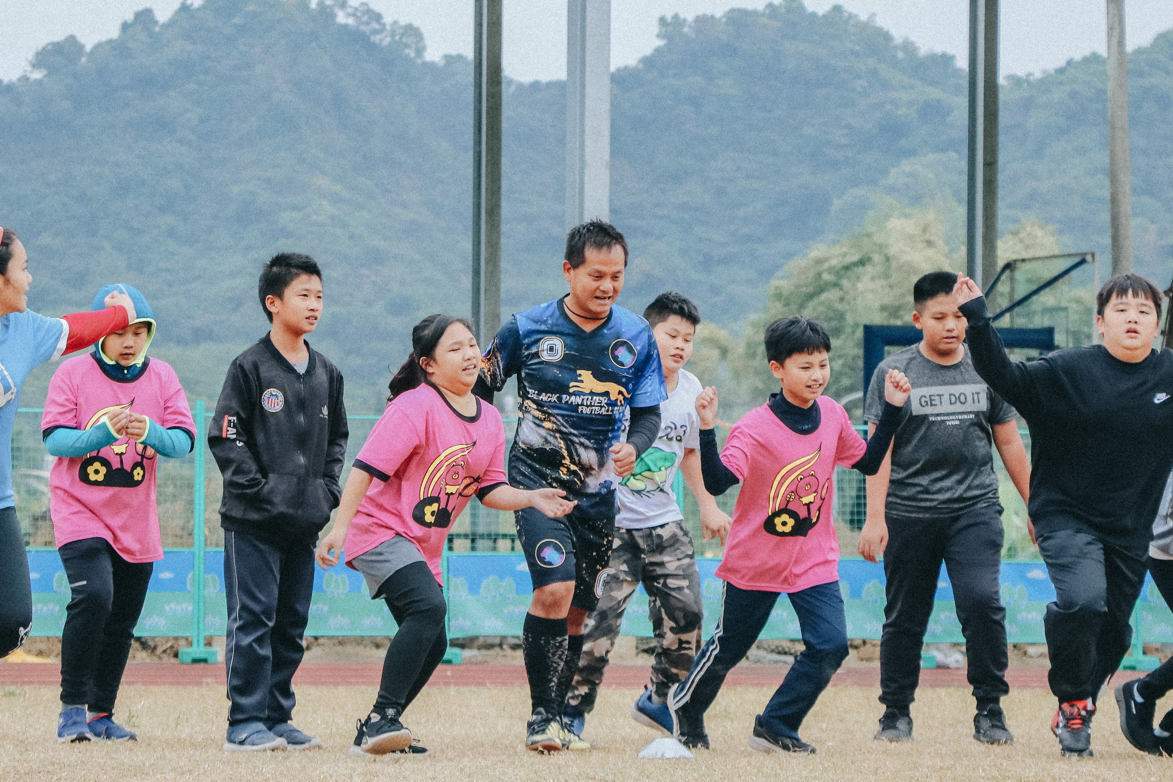 一群小學生在運動場上進行跑步活動。孩子們穿著粉紅色和黑色運動服，在紅色跑道上奔跑，背景是綠色山林和體育設施。場面充滿朝氣，展現學校體育運動的活力。