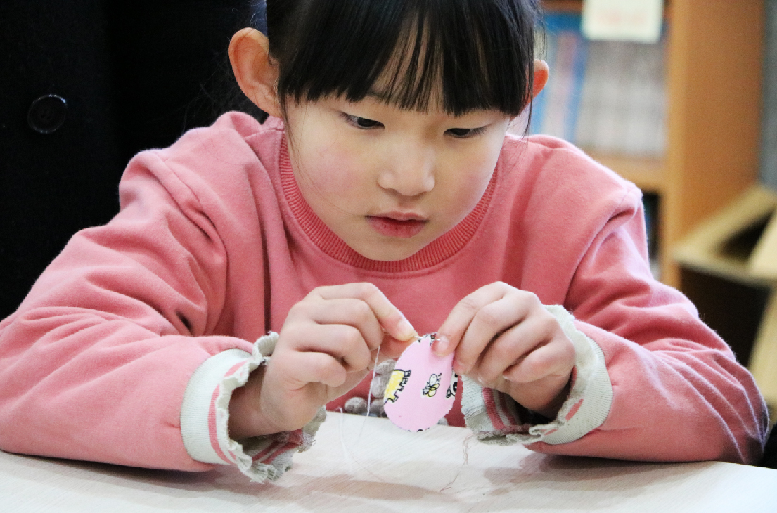 一位穿著粉紅色上衣的年幼女童，低頭專注地用雙手操作一個粉紅色的小物件，放在白色工作台上。她的神情認真投入，專注於手邊的活動，背景為室內教室環境。