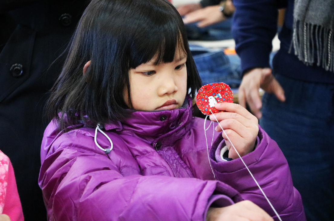 一位穿著紫色羽絨外套、留著齊肩黑髮的小女孩，低頭專注地看著手中的紅色閃亮飾品。背景有其他穿著深色衣物的人，暗示這是一場室內活動或聚集場景。